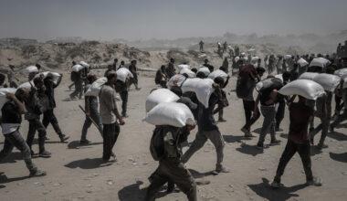 Palestinians carry white large sacks of aid supplies on their backs whilst walking across a dusty road in Beit Lahia, northern Gaza 