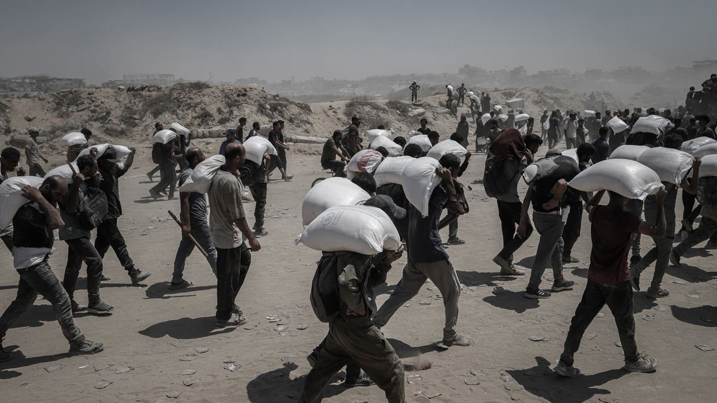 Palestinians carry white large sacks of aid supplies on their backs whilst walking across a dusty road in Beit Lahia, northern Gaza 