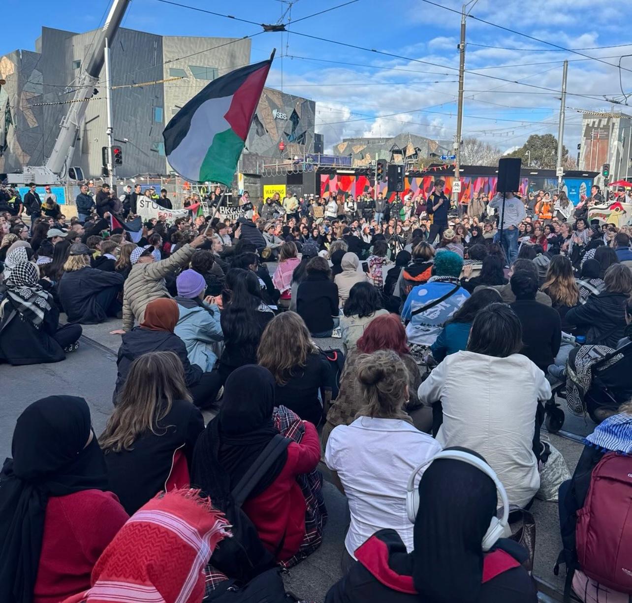 A large group of people are sitting on a street with some holding Palestinian flags during a protest.