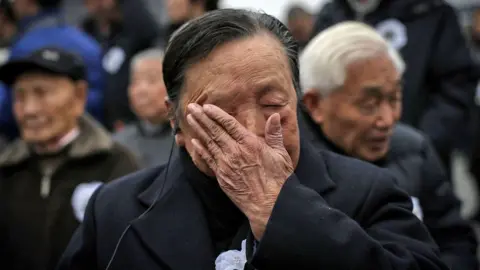STR/AFP via Getty Images An elderly man wearing a suit with an ear piece in his ear wipes tears off his face. Behind him more elderly men can be seen, slightly blurred