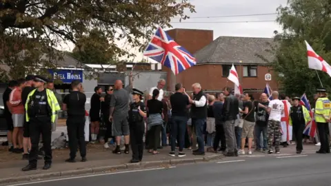 A small crowd of protesters holding Great Britain flags stand on the pavement outside a hotel as three police officers stand nearby.