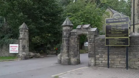 BBC A grand entrance to a park with a sign to the right that reads: Manor Heath Car Park. There are trees and stone buildings and a road leading between stone pillars.