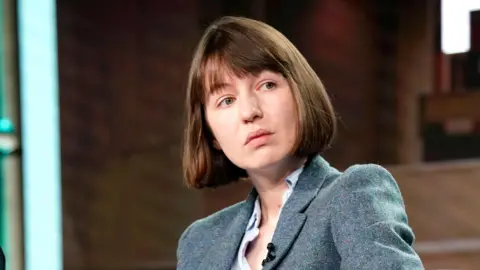 Getty Images Sally Rooney, who has short brown hair and a fringe, and is wearing a woollen suit jacket and a shirt, listens to someone speak. 