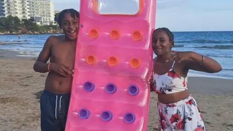 Family Ameiya (R) and Ricardo Del Brocco (L) on holiday. They are either side of an pink inflatable. They are on a beach and smiling.  Ameiya has short dark hair and wears a two piece costume of white with a floral design. Ricardo has short dark hair in braids and wears a blue pair of shorts. 