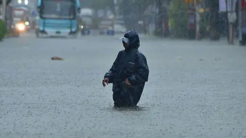 Hindustan Times via Getty Images A man wearing a black raincoat wades through knee-deep water in Mumbai, Maharashtra.