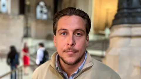 A man with brown hair. He is wearing a pale blue shirt and beige jumper. 