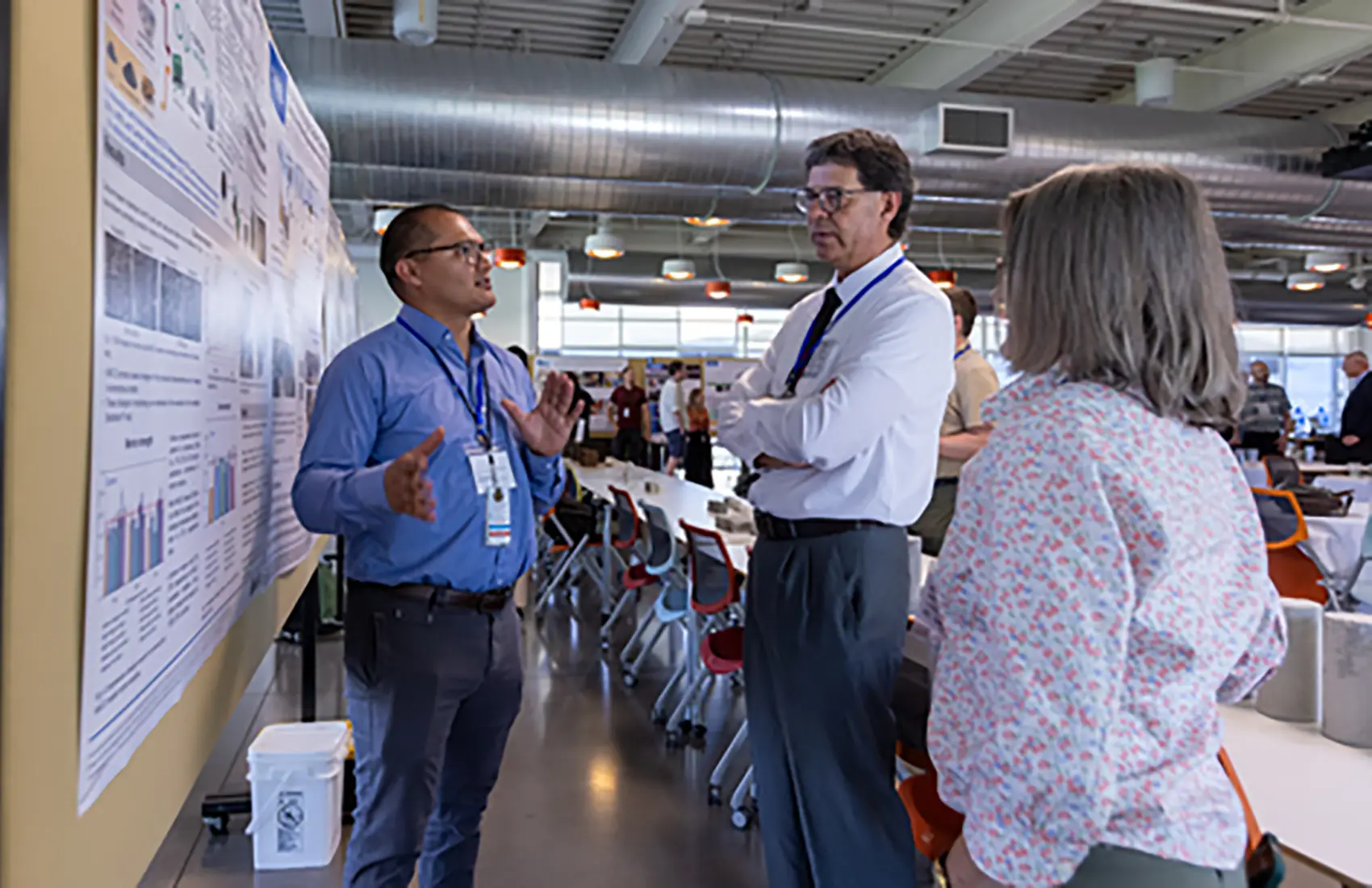 An individual presents in front of a poster while two other individuals listen.