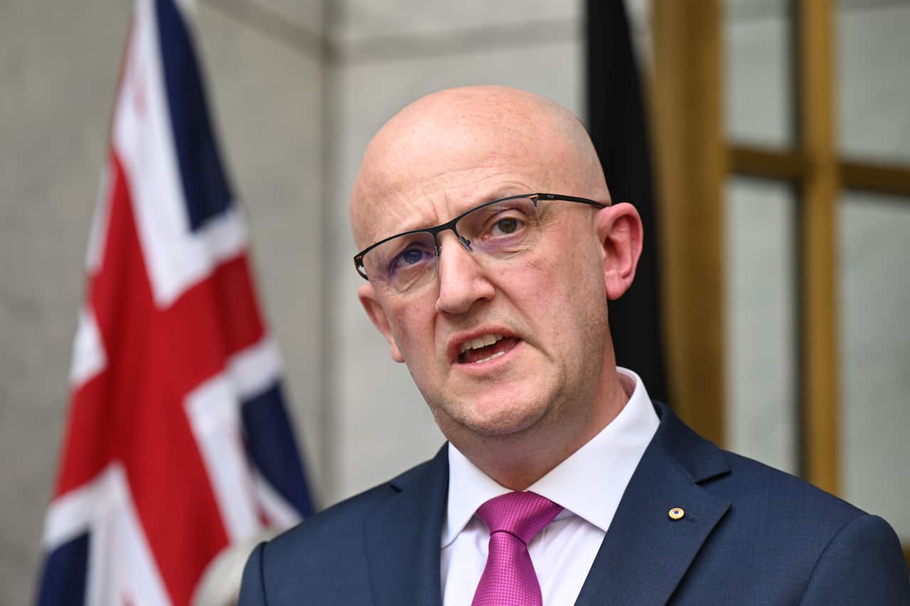 A man in a suit is speaking in front of the Australian flag.