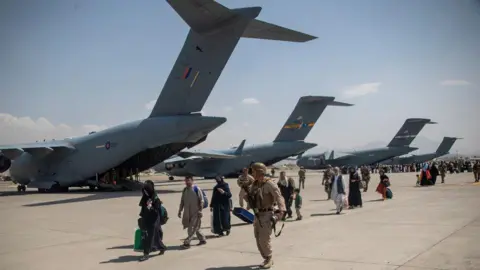 MOD/Crown Copyright/2021 Members of the UK Armed Forces leading Afghan refugees past a row of RAF transport planes at Kabul airport. The refugees' faces have been blurred