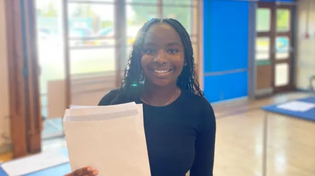 Girl standing in gymnasium smiles at the camera while holding exam envelope in hand