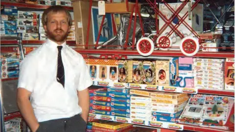 The Entertainer Gary Grant pictured in 1981 standing inside his first toy shop surrounded by board games wearing a white shirt with a dark tie tucked into his buttons