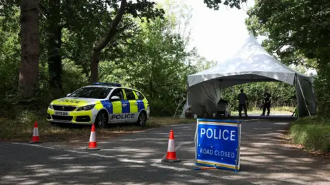 Reuters A police car is parked on the side of a road, blocked by traffic cones and a sign reading "Police road closed", in the hamlet of Dean in the Cotswolds. Behind there is a white tent, a police officer and a security patrol.