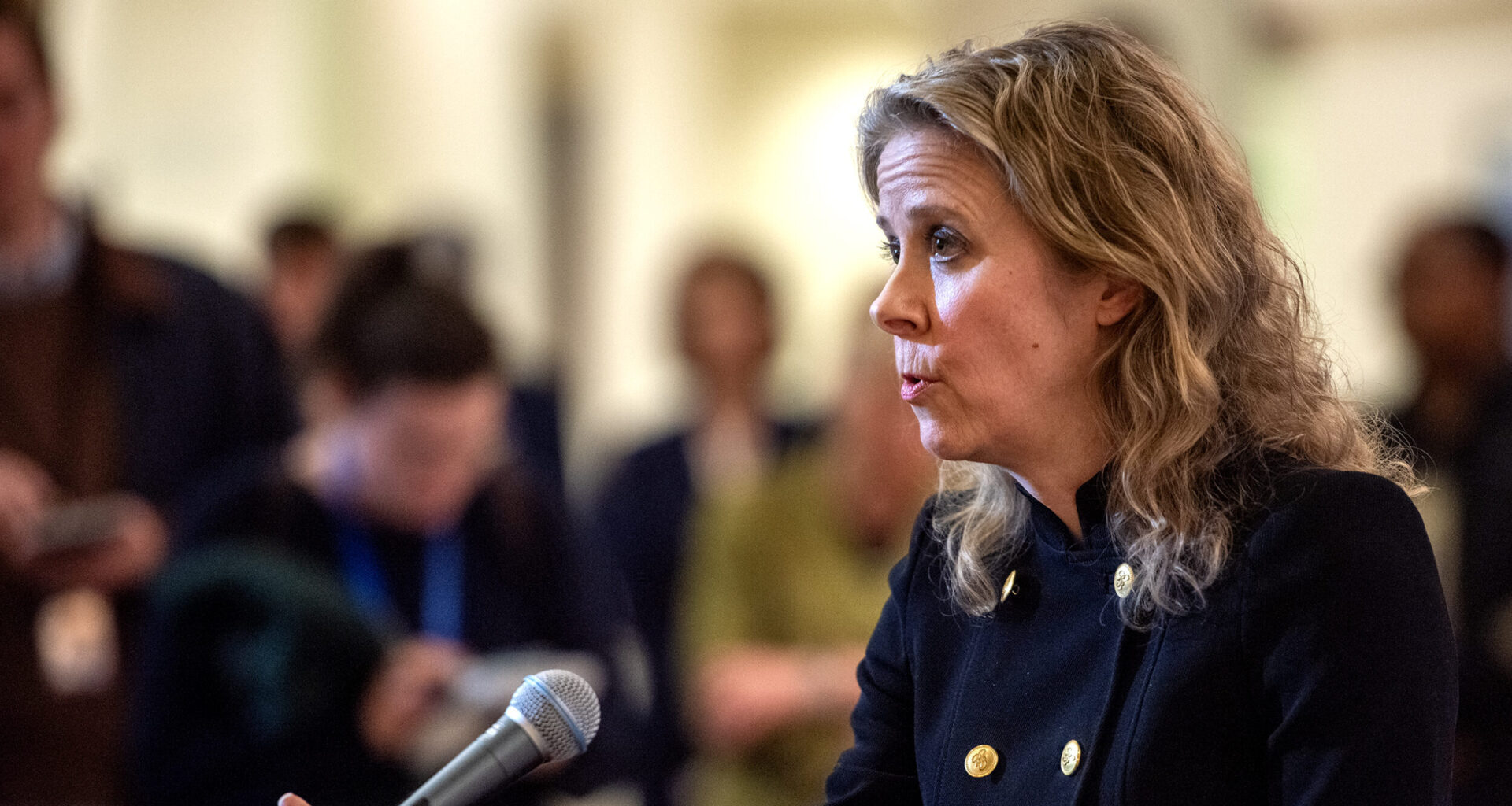 Woman speaking at a podium with a microphone, surrounded by an audience.