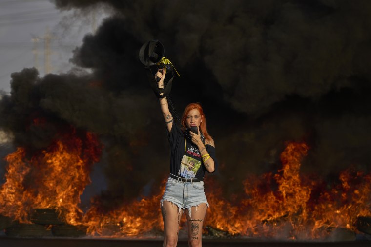 An activist blocks a highway during a protest, standing in front of a fire.