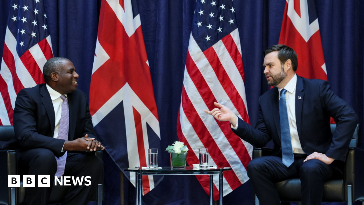 David Lammy smiles across a table at JD Vance, who is talking and gesticulating at a meeting. The backdrop is large UK and US flags.