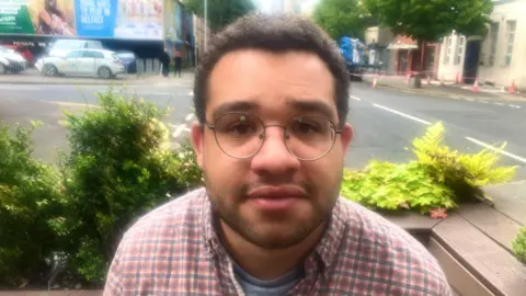 Christian Nsonwu is looking directly at the camera with a neutral expression on his face. He is sitting on a wooden bench with some shubbery in the background, beside a main road in Belfast city centre. Mr Nsonwu has a pair of thin wired circular glasses, short dark hair and is wearing a red, blue and white flannel shirt.