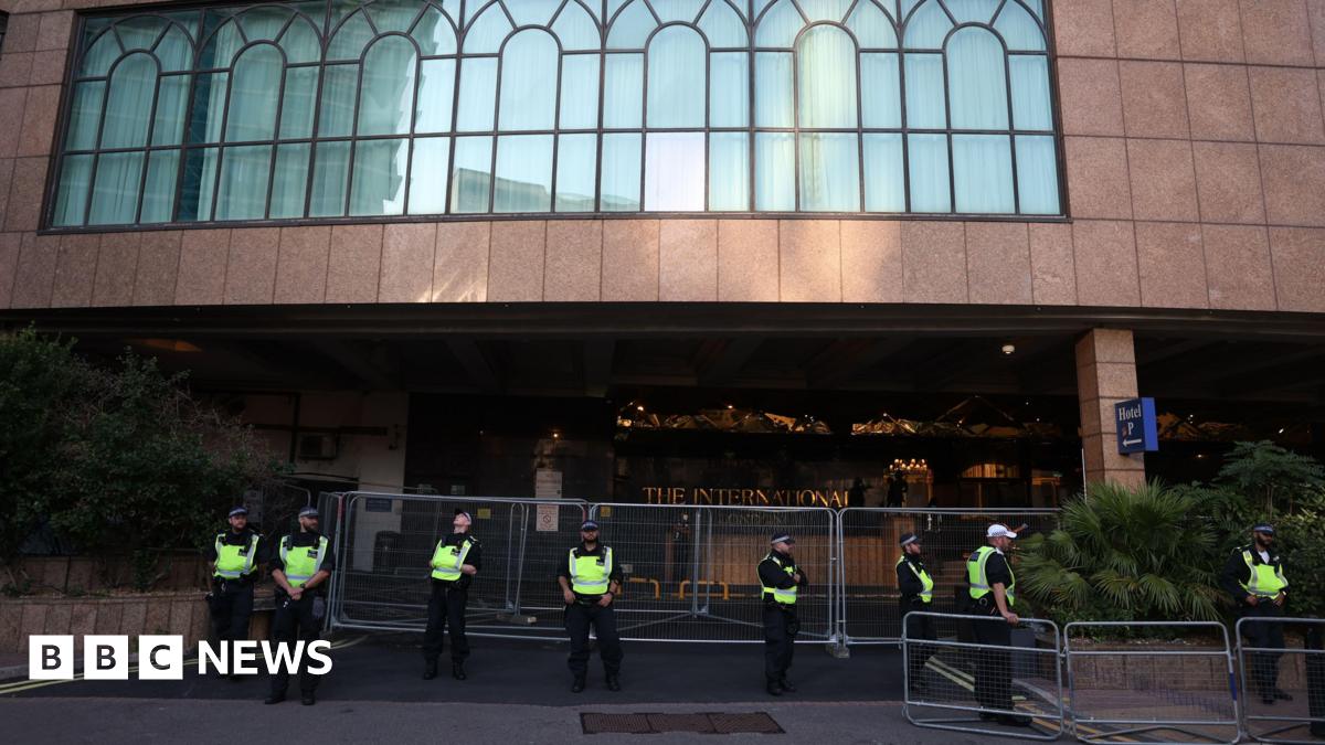 Police officers in high-visibility jackets stand in a line behind metal barriers outside the International Hotel in east London, with the building’s large arched windows visible above the entrance.