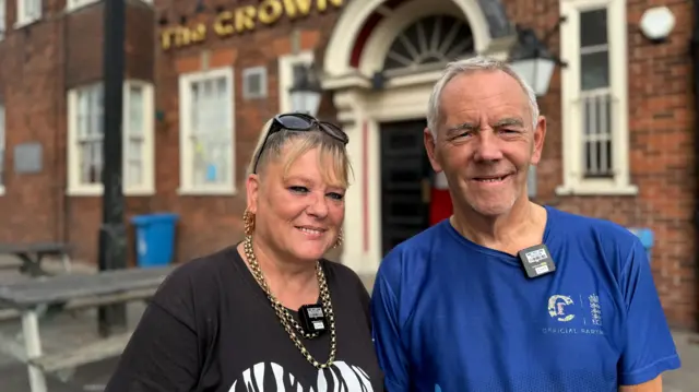 Two people stand outside a brick building with a sign reading "THE CROWN". The building has an arched entrance, multiple windows and picnic tables. A woman on the left wears a black zebra-print shirt with sunglasses on her head. A man on the right is wearing a blue shirt with white text.