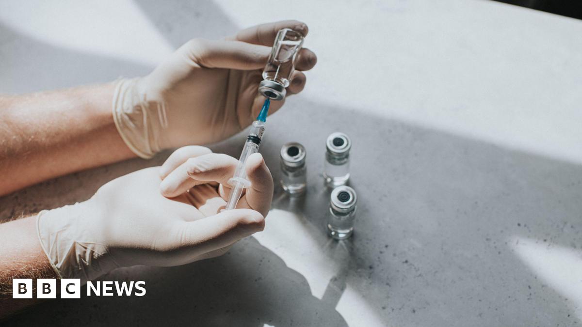A gloved medical professionals' hand uses a syringe to extract liquid from some vials on a table.