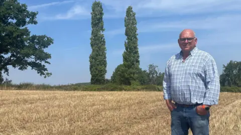 BBC/Malcolm Prior Martin, dressed in a light-blue-checked shirt and blue jeans, stands with his hands in his pockets in a wheat field that has been harvested. He is bald and wearing rectangular glasses. A blue sky with little cloud indicates it is still hot while the yellow crop field appears dry. On the horizon are green trees.