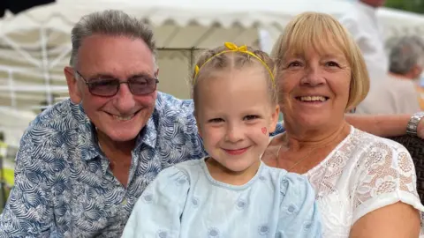 Avril Hackett and her husband with their granddaughter Emily in between them. She is holding a colourful teddy and there is a large, white marquee-style tent behind them