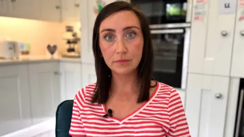 Niamh Hagan has shoulder-length dark brown hair and is wearing a striped red and white top. She is sitting in a kitchen, behind her white kitchen units and an oven built into the wall can be seen.