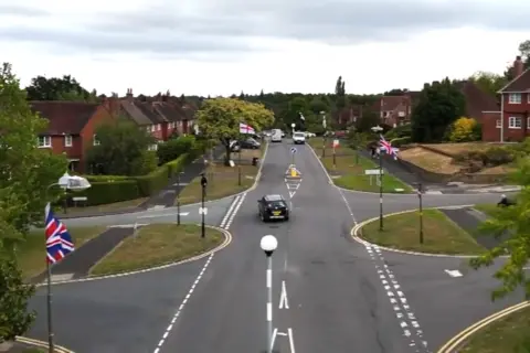 Union Jack and St George flags hang from every lamppost on a street in Birmingham, seen from an aerial drone shot.