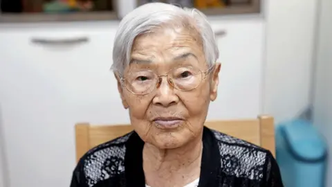 BBC/Hyojung Kim An old woman white short white hair and wire frame glasses sits in a chair and looks into the camera
