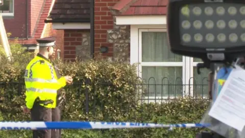 A police officer is stood near a fenced-off front garden of a house. There is also blue and white police tape.