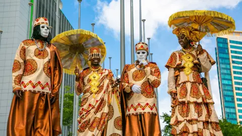 EPA Four people stand in a row, wearing African print outfits. Two wear white masks, the other two carry parasol