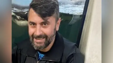 Essex Police Cumali Turhan has black hair swept to one side and a greying, black beard. He is wearing a black jacket and appears to be sitting in front of an aquarium display