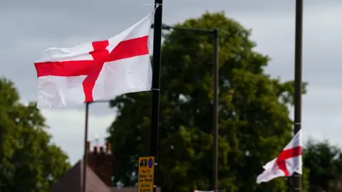 PA Media Two red and white St George's flags fly in the wind, having been tied to lampposts on a residential street.