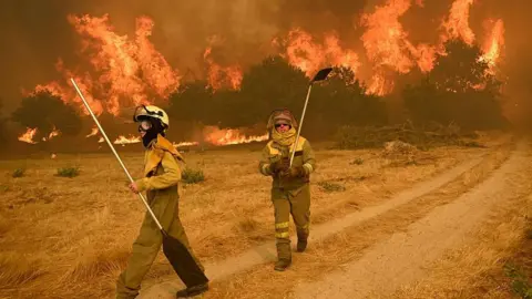 Getty Images Two female firefighters tackle a raging inferno in a Spanish woodland scene. The grass around them is dry and parched, and the trees in the background are completely aflame.