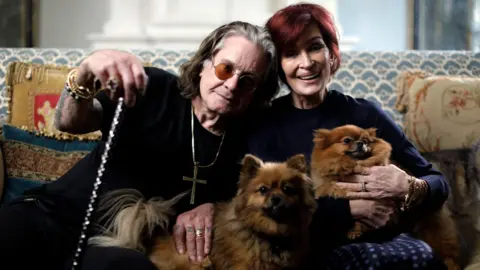 Ruaridh Connellan/Expectation/BBC Ozzy Osbourne, an elderly man with long grey/black hair, a crucifix around his neck, sits on a sofa while holding a cane. He is leaning on his wife Sharon - a woman with short red hair and wearing blue. They are holding two small brown dogs.