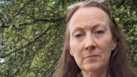 Helen Hickman Helen, a small business owner, stands by a tree with her long brown hair blowing in the wind.