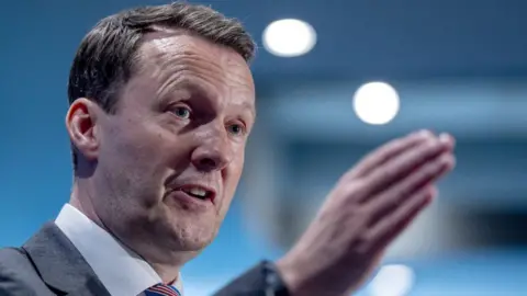 PA Media A man with dark hair speaks with his right hand held in front of his face. He is wearing a grey suit and white shirt. 