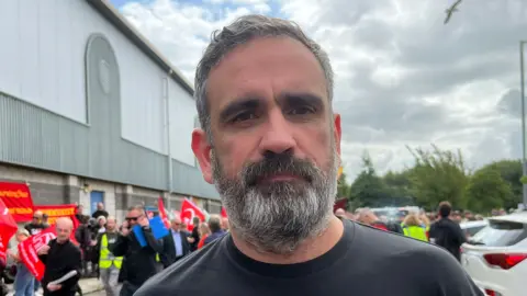 BBC A man with greying hair and a beard, wearing a black T shirt with a demonstration in the background