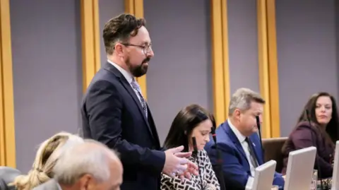 Senedd Cymru Hefin David in the Senedd. He has a brown beard and brown short hair. He wears black square glasses.