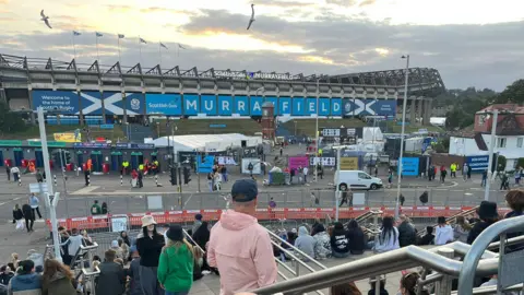 A group of Oasis fans outside Murrayfield Stadium sitting on the steps of the tram stop with the stadium in the background. 