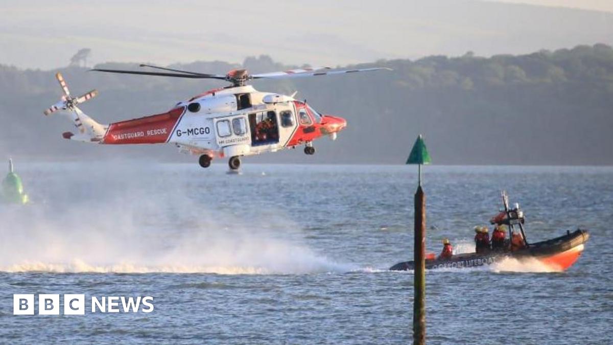 A coastguard helicopter flies low over the sea with a lifeboat slightly in front of it. A wooded shoreline can be seen in the background.