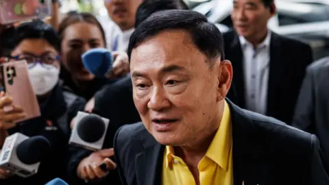 Getty Images Side view of Thaksin in a black blazer and yellow shirt, surrounded by reporters who are holding microphones out to him.