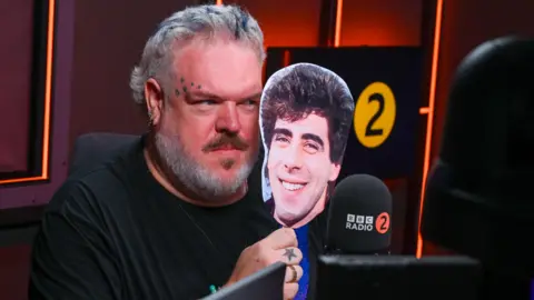 Kristian Nairn looking out from behing a mask of Gary Davies's face in the radio studio with Radio 2 logos