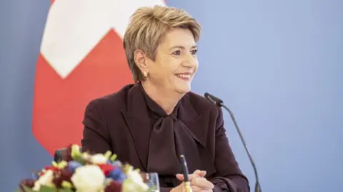 Getty Images Switzerland's president Karin Keller-Sutter smiling sat at a desk with a blue background and part of a swiss flag behind her