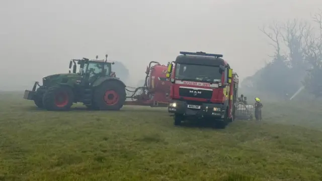 A tractor stands alongside a fire engine, engulfed by thick smoke