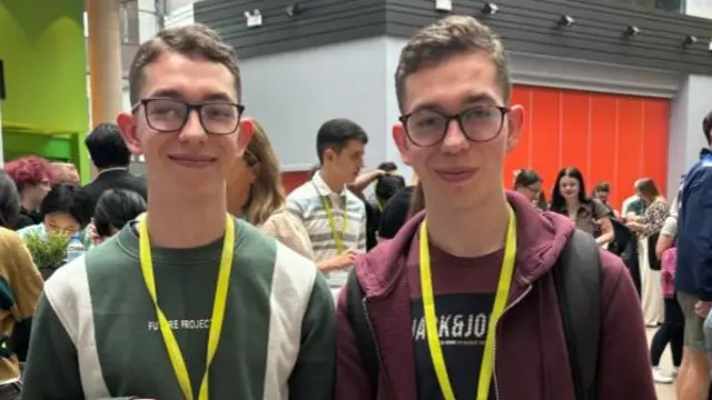 Twins smiling at the camera as both hold their results papers in a busy room