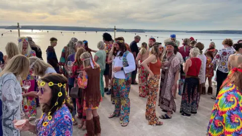Mike Conner A large group of people stand on a flat concrete surface, with the sea stretching beyond then. They are all wearing colourful 60s themed fancy dress costumes, with many wearing different coloured round sunglasses, long wigs, and headbands.  