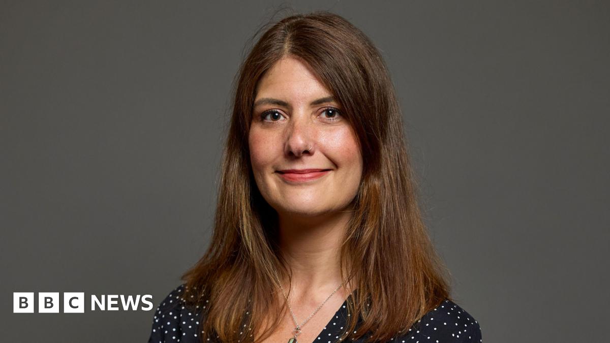 MP Cat Eccles, a woman with brown hair, is stood looking at the camera and smiling. She is wearing a necklace and a black top with white dots.