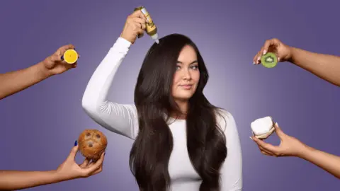Hair Syrup A young woman wearing a plain white, long-sleeved top. She has long, dark wavy hair that goes to her stomach and she is holding a hair serum bottle in her right hand, above her head, pretending to pour it on herself. She is in front of a plain purple background and four arms are holding food items in her direction, including a coconut, lime and a kiwi 