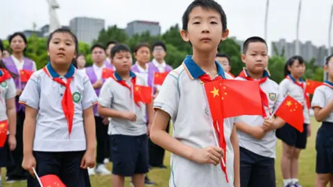 CFOTO/Future Publishing via Getty Images A school boy holds a Chinese flag up in his hand. Behind him are lots of school children in white tops and blue shorts with red and blue and white neckerchiefs. they are standing on a field with tees and high rises in the background