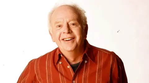 BBC Ray Brooks, a man with white hair wearing a striped red button-down shirt over a black t-shirt, smiles at the camera in front of a white background.
